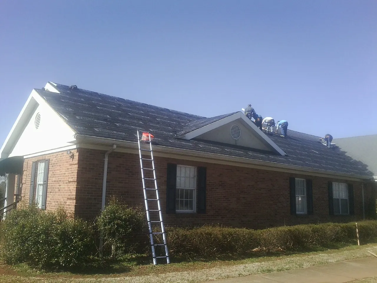 Crew tearing shingles off a brick ranch roof with a ladder set — Emergency Roof Repair work by our crew in Clontarf