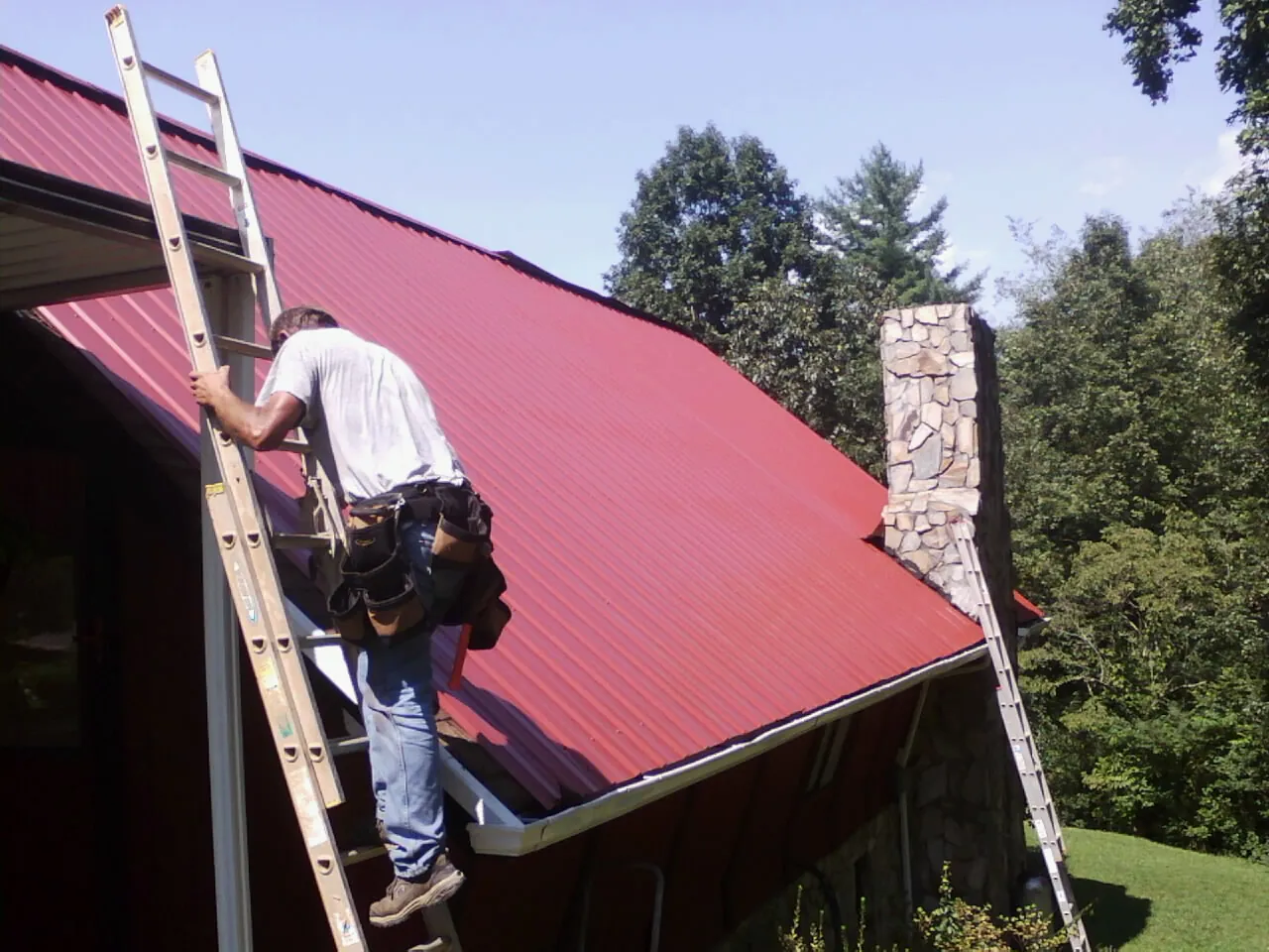 Roofer on a ladder beside a red metal roof and stone chimney — Soffit & Fascia Repair by Roofing Brothers in Clontarf